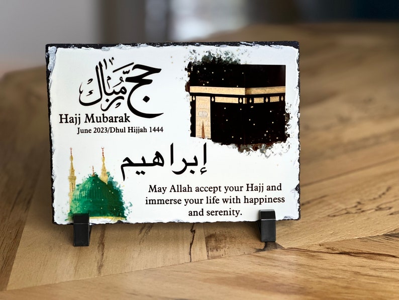 May include: White slate plaque with a black and gold image of the Kaaba, a green mosque, and Arabic calligraphy. The text reads "Hajj Mubarak June 2023/Dhul Hijjah 1444" and "May Allah accept your Hajj and immerse your life with happiness and serenity."