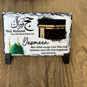May include: A black slate plaque with a white background featuring an image of the Kaaba, a mosque, and Arabic calligraphy. The text reads "Hajj Mubarak June 2023/Dhul Hijjah 1444" and "Jasmeen May Allah accept your Hajj and immerse your life with happiness and serenity."