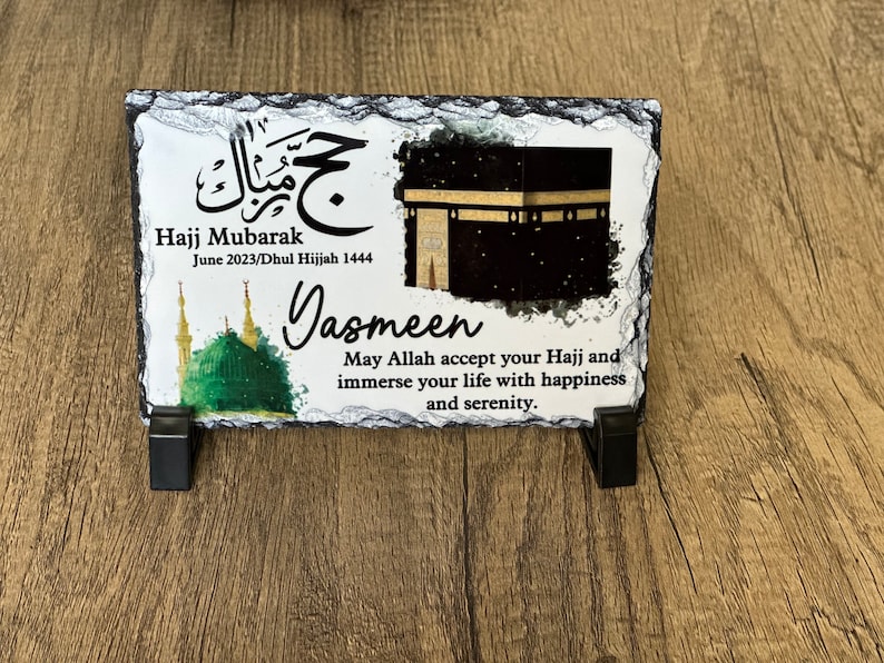 May include: A black slate plaque with a white background featuring an image of the Kaaba, a mosque, and Arabic calligraphy. The text reads "Hajj Mubarak June 2023/Dhul Hijjah 1444" and "Jasmeen May Allah accept your Hajj and immerse your life with happiness and serenity."