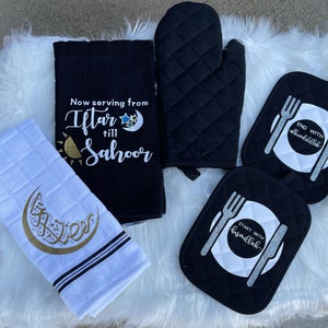 May include: A set of kitchen textiles featuring a Ramadan theme. The set includes a black kitchen towel with the text "Now serving from Iftar till Sahor", a white kitchen towel with a crescent moon and Arabic calligraphy, a black oven mitt, and two black pot holders. One pot holder has the text "Start with Bismillah" and the other has the text "End with Alhamdulillah".