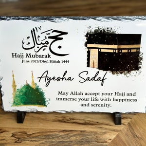 May include: A white slate plaque with a black and gold illustration of the Kaaba, a green mosque, and Arabic calligraphy. The text reads "Hajj Mubarak June 2023/Dhul Hijjah 1444 Ayesha Sadaf May Allah accept your Hajj and immerse your life with happiness and serenity."