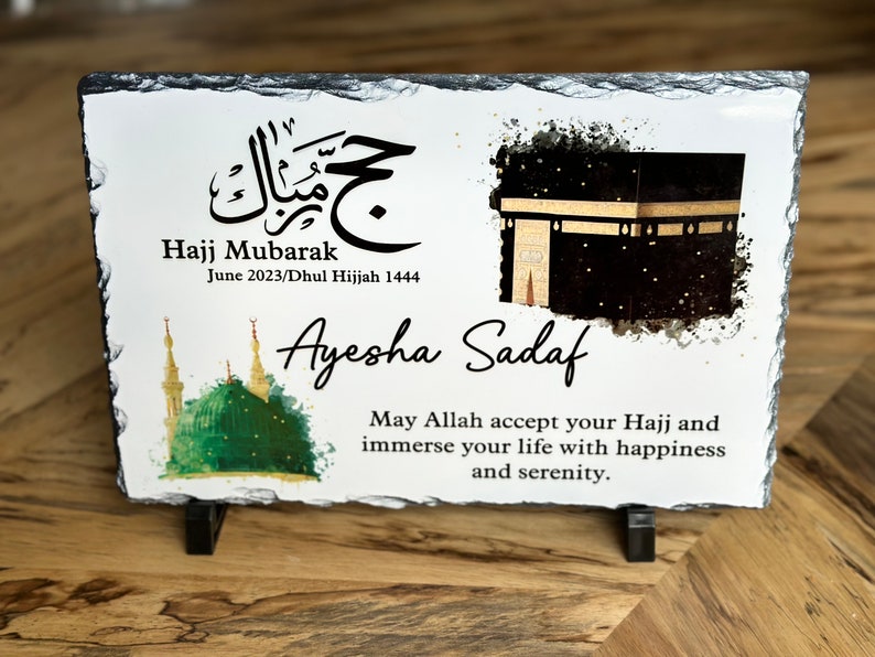 May include: A white slate plaque with a black and gold illustration of the Kaaba, a green mosque, and Arabic calligraphy. The text reads "Hajj Mubarak June 2023/Dhul Hijjah 1444 Ayesha Sadaf May Allah accept your Hajj and immerse your life with happiness and serenity."