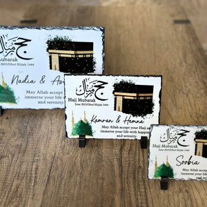 May include: Three personalized slate plaques with a black and white image of the Kaaba, a green mosque, and Arabic calligraphy. The text on the plaques reads "Hajj Mubarak" and includes the names Nadia & As, Kamran & Hemna, and Soobia. Each plaque has a message wishing the person a happy Hajj and a peaceful life.