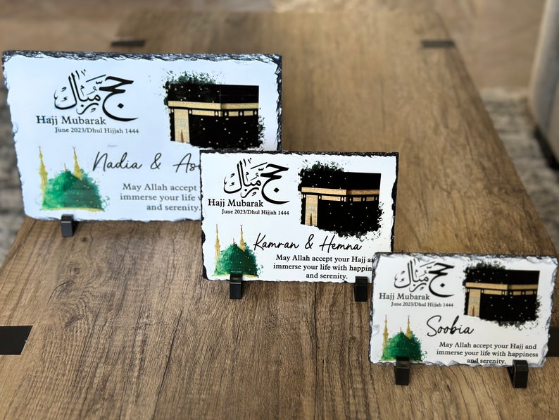 May include: Three personalized slate plaques with a black and white image of the Kaaba, a green mosque, and Arabic calligraphy. The text on the plaques reads "Hajj Mubarak" and includes the names Nadia & As, Kamran & Hemna, and Soobia. Each plaque has a message wishing the person a happy Hajj and a peaceful life.