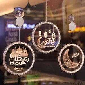 May include: Three round, hanging, LED light decorations with a Ramadan theme. The decorations feature a crescent moon, a mosque, and Arabic calligraphy. Each decoration has a suction cup for hanging.