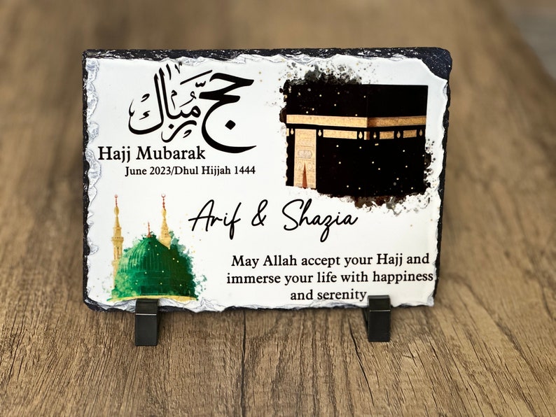 May include: A white slate plaque with a black and gold image of the Kaaba, a green mosque, and Arabic calligraphy. The text reads "Hajj Mubarak June 2023/Dhul Hijjah 1444 Arif & Shazia May Allah accept your Hajj and immerse your life with happiness and serenity."
