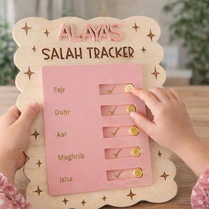 May include: A wooden Salah tracker with the name "ALAYA'S" at the top. The tracker has a pink insert with slots for prayer times and gold-colored tokens. The frame is light-colored wood with star accents.