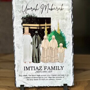 May include: A black slate plaque with a watercolor illustration of the Kaaba in Mecca, Saudi Arabia. The plaque has the text "Umrah Mubarak" and "IMTIAZ FAMILY 2023 1445 AH" and a message about the serenity of the holy lands.