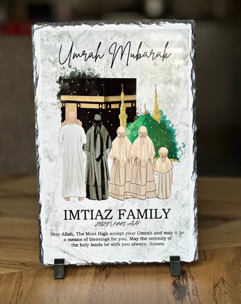 May include: A black slate plaque with a watercolor illustration of the Kaaba in Mecca, Saudi Arabia. The plaque has the text "Umrah Mubarak" and "IMTIAZ FAMILY 2023 1445 AH" and a message about the serenity of the holy lands.