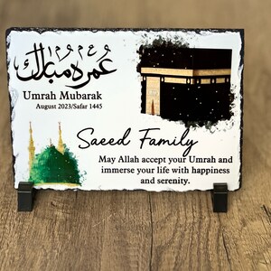 May include: A white slate plaque with a black and gold image of the Kaaba, a green mosque, and Arabic calligraphy. The text reads "Umrah Mubarak August 2023/Safar 1445 Saeed Family May Allah accept your Umrah and immerse your life with happiness and serenity."