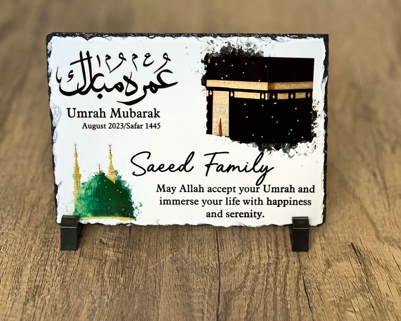 May include: A white slate plaque with a black and gold image of the Kaaba, a green mosque, and Arabic calligraphy. The text reads "Umrah Mubarak August 2023/Safar 1445 Saeed Family May Allah accept your Umrah and immerse your life with happiness and serenity."