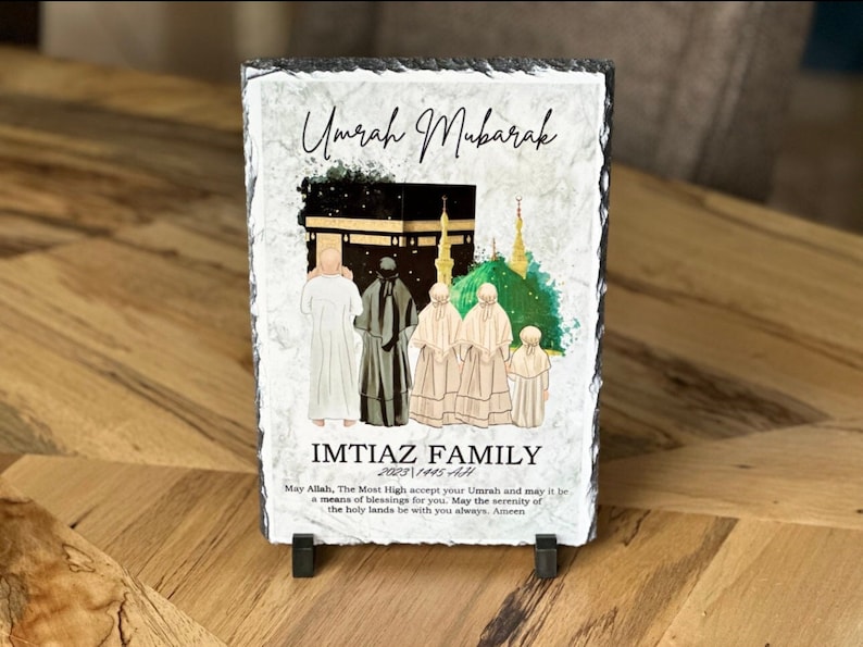 May include: A black slate plaque with a watercolor illustration of the Kaaba and a family in white robes. The text "Umrah Mubarak" and "IMTIAZ FAMILY 2023 1445 AH" is printed on the plaque. The plaque is on a wooden surface.