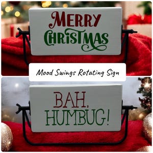 May include: A rectangular white rotating sign held by a black metal frame. One side features the red and green text Merry Christmas in a decorative script. The other side displays the red and green text BAH, HUMBUG! in a bold block font.