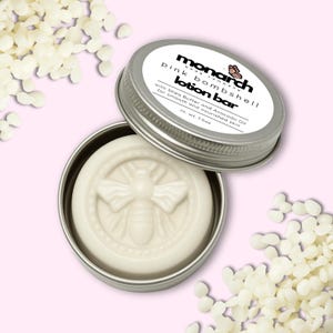 May include: A white lotion bar in a silver tin, with the text "pink bombshell lotion bar" and "Monarch Body Company". The lotion bar has a bee design. The tin is open, revealing the bar. Scattered white beads surround the tin.