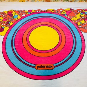 May include: A colorful, retro-style textile featuring a large, concentric circle design. The center is yellow, surrounded by orange, pink, and blue rings. The design includes the text "peter max". The background is white.