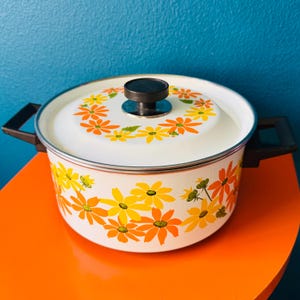 May include: White enamel pot with a black knob on the lid. The pot has a floral pattern with orange and yellow flowers on a white background.