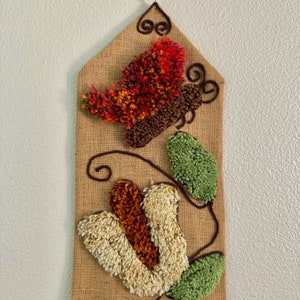 May include: A wall hanging made of burlap with a brown, red, orange, and white yarn butterfly and green yarn leaves. The butterfly is in the upper left corner of the burlap and the leaves are in the lower right corner.