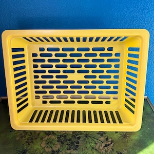 May include: A rectangular yellow plastic storage basket featuring a grid-like pattern of horizontal slots on the sides and base. The basket has a wide, flat rim and an open top design, providing a structured container for organizing various items.