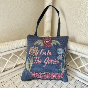 May include: A blue and green floral pillow with a loop handle. The pillow has the text "I'm in the Garden" embroidered on it.