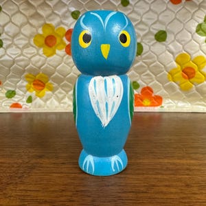 May include: A blue wooden owl figurine featuring yellow eyes with black pupils, a yellow beak, and a white painted chest detail. The owl has stylized white markings around the eyes and light blue claw designs at the base, standing in an upright position.