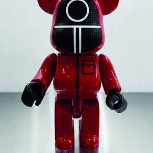 May include: A glossy red bear-shaped collectible figure with a black mask featuring a white circle and vertical line design. The figure has black hands and a black vertical zipper detail on the chest, resembling a jumpsuit with a square pocket.