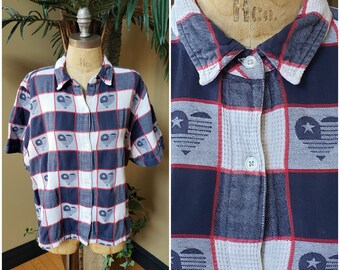 Puede incluir: Una camisa de botones a cuadros azules y blancos con ribetes rojos y un diseño de bandera estadounidense en forma de corazón. La camisa tiene mangas cortas y cuello.