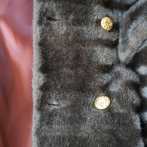 Puede incluir: Primer plano de una chaqueta de piel sint&eacute;tica marr&oacute;n oscuro con tres botones dorados. Los botones tienen un dise&ntilde;o decorativo.