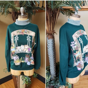 May include: A green sweatshirt with a colorful illustration of two kittens on a swing. The kittens are surrounded by flowers, baskets, and other garden items. The sweatshirt has a white and green checkered turtleneck.