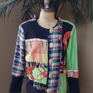 May include: A patchwork jacket with a unique design. It features a mix of plaid, floral, and solid fabric panels in various colors, including green, red, and black. The jacket has decorative buttons and fringe accents, creating a bold and artistic look.
