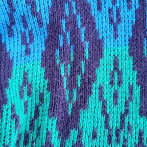 May include: Close-up of a knitted fabric with a blue, green, and purple pattern. The fabric is textured and has a fuzzy appearance.