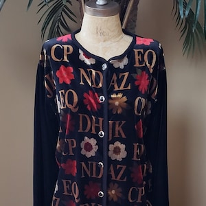 May include: Black velvet cardigan with a button-up front. The cardigan is covered in a pattern of gold letters and red and white flowers.