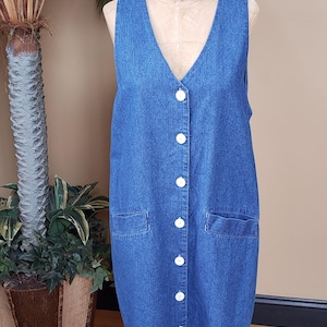 May include: A sleeveless, knee-length denim dress with a V-neckline and white buttons down the front. The dress features two front pockets and is displayed on a mannequin. The dress is a medium blue color.