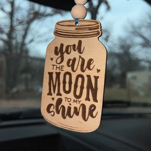 May include: Wooden car air freshener shaped like a mason jar with the text "you are the moon to my shine" in a brown font.
