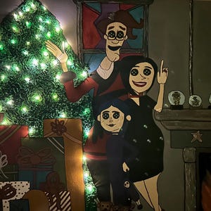 Op de afbeelding: Een grillig kerstkunstwerk met een familie met knoogogen, een versierde kerstboom met lichtjes en ingepakte cadeaus. Het kunstwerk heeft een open haard en een ronde spiegel met een stel.