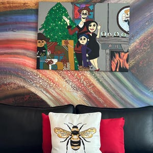 Op de afbeelding: Een schilderij met een familiesc&egrave;ne met een kerstboom en cadeaus. Daaronder ligt een wit kussen met een gedetailleerd geborduurd bijenontwerp op een zwarte leren bank, vergezeld van een rood kussen.
