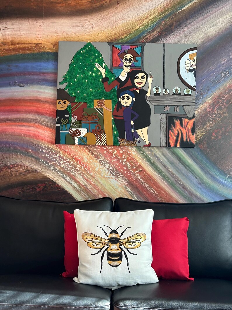 Op de afbeelding: Een schilderij met een familiesc&egrave;ne met een kerstboom en cadeaus. Daaronder ligt een wit kussen met een gedetailleerd geborduurd bijenontwerp op een zwarte leren bank, vergezeld van een rood kussen.