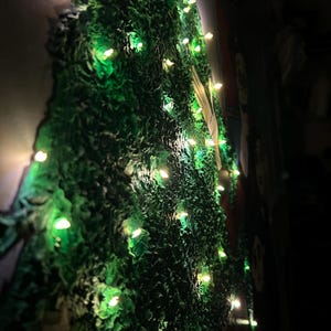 Op de afbeelding: Een groene muur met textuur en ingebouwde lichten. De muur heeft een ruw, ongelijkmatig oppervlak en de lichten zijn klein en gelijkmatig verdeeld, waardoor een zachte, warme gloed ontstaat. Het algehele effect is een decoratieve, verlichte display.