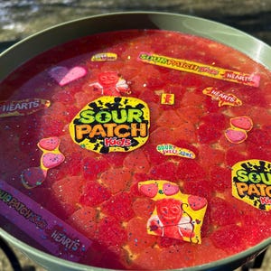 May include: A green bowl filled with red liquid and various Sour Patch Kids candies. The candies include heart-shaped gummies, Sour Patch Kids logos, and the text "HEARTS" and "SOUR PATCH Kids HEARTS".