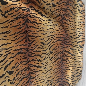 Può includere: Un tessuto tessile caratterizzato da un motivo ripetitivo a strisce di tigre nei toni del giallo dorato, arancione e nero. Il materiale presenta una consistenza superficiale morbida e leggermente riflettente con pieghe e drappeggi.