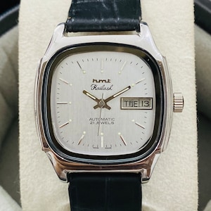 Op de afbeelding: Een zilverkleurig polshorloge met een witte wijzerplaat en een zwarte leren band. De wijzerplaat van het horloge heeft een datumscherm dat "TUE 13" weergeeft en de tekst "HMT Kailash AUTOMATIC 21 JEWELS INDIA MADE".