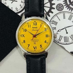 May include: A yellow analog wristwatch with a silver case and black leather band. The watch face has black Roman numeral hour markers and a red second hand. The watch face also has the text "humut Janata" and "PARA SHOCK 17 JEWELS".
