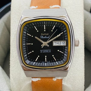Op de afbeelding: Een zilveren en gouden herenhorloge met een zwarte wijzerplaat, een bruin leren bandje en de woorden "HMT Kailash Automatic 21 Jewels" gedrukt op de wijzerplaat. Het datumscherm toont "THU 12".