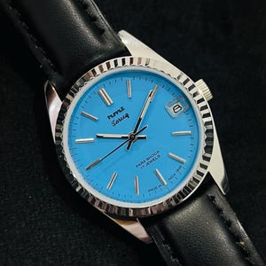 Op de afbeelding: Een zilveren polshorloge met een blauwe wijzerplaat en een zwart leren bandje. De wijzerplaat van het horloge heeft witte markeringen en de tekst "HMT Tareeq Para Shock 17 Jewels Made in India" rond de rand.