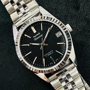 Op de afbeelding: Een zilveren polshorloge met een zwarte wijzerplaat en zilveren wijzers. Het horloge heeft een zilveren band en op de wijzerplaat staat "HMT Tareef Para Shock 17 Jewels".