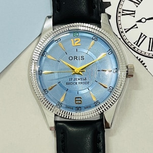Peut inclure: Une montre-bracelet argentée avec un cadran bleu et des accents dorés. La montre a un bracelet en cuir noir et indique "ORIS" et "17 JEWELS SHOCK PROOF" sur le cadran.