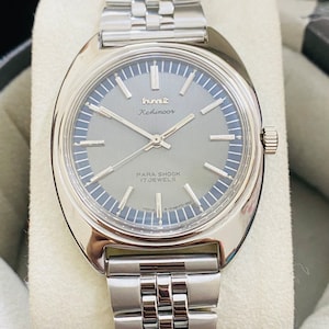 May include: A silver wristwatch with a silver metal strap. The watch face is grey with silver hands and markers. The watch face says "HMT Kohinoor Para Shock 17 Jewels".