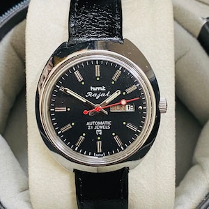 Op de afbeelding: Een polshorloge met een zwarte wijzerplaat, zilveren wijzers en markeringen. De wijzerplaat van het horloge heeft de tekst "HMT Rajal Automatic 21 Jewels" en een datumscherm met de weergave "SUN 19". Het horloge is voorzien van een zwarte leren band.