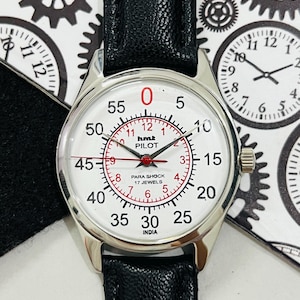 May include: A silver wristwatch with a white face and black leather strap. The watch face has red markings and the text "hmt PILOT PARA SHOCK 17 JEWELS INDIA".