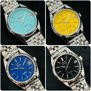 Op de afbeelding: Vier zilveren polshorloges met verschillende gekleurde wijzerplaten. De horloges hebben zilveren banden en een zilveren kast. De wijzerplaten zijn blauw, zwart, geel en lichtblauw. Op elk wijzerplaat staat de tekst "Humt Loreeg" en "Para Shock 17 Jewels" gedrukt.