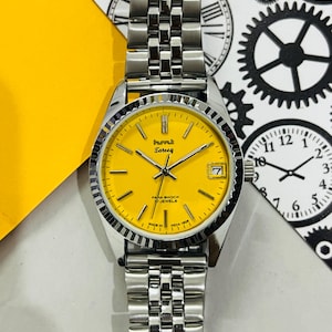 Op de afbeelding: Een zilveren polshorloge met een gele wijzerplaat en een zilveren metalen band. De wijzerplaat van het horloge heeft zwarte markeringen en de tekst "Purvuk Tareeq Para Shock 17 Jewels Made in India 1809".